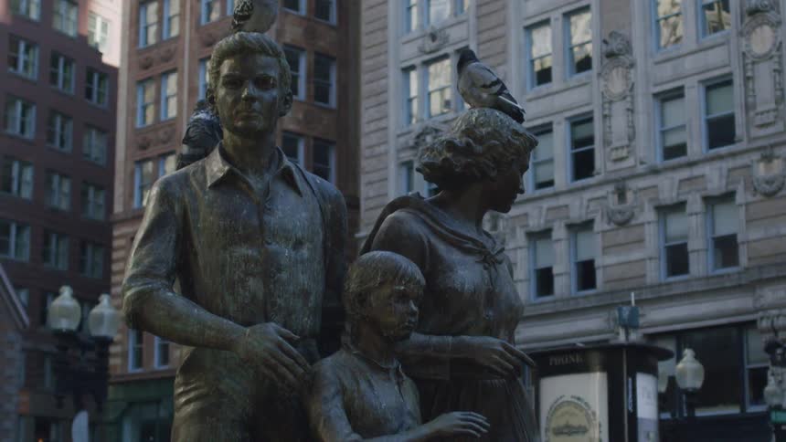 Pigeons are perched on the heads and shoulders of a bronze statue depicting a man woman and childthe statues are positioned in an urban setting with large multistory buildings and a phone booth in the background