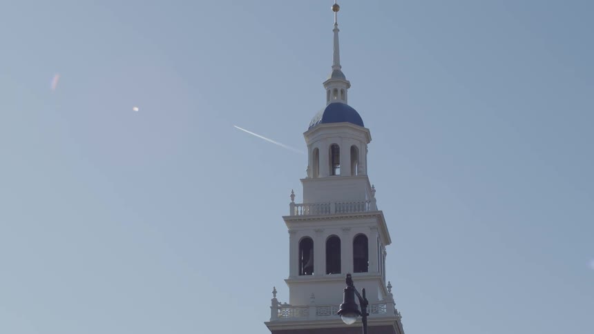 The video shows a white multitiered steeple with a blue dome and a golden finial set against a clear sky a faint white vapor trail from an airplane stretches diagonally across the blue background behind the architectural structure