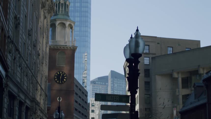 As the camera tilts upward from a bustling downtown street a historic brick clock tower is revealed standing in sharp contrast to modern glass skyscrapers while a flock of pigeons takes flight from a nearby building