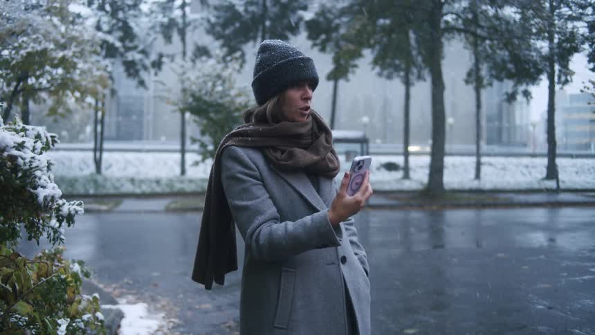 A woman bundled in a warm winter coat talks to her smartphone as soft snowflakes drift down around her on a quiet city street