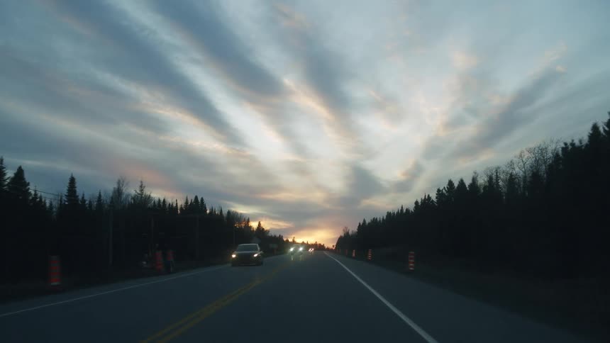 Traveling along a forested highway at dusk a vehicle heads toward a vibrant golden sunset as wispy clouds stretch across the vast sky and oncoming cars with glowing headlights pass by dense silhouettes of evergreen trees