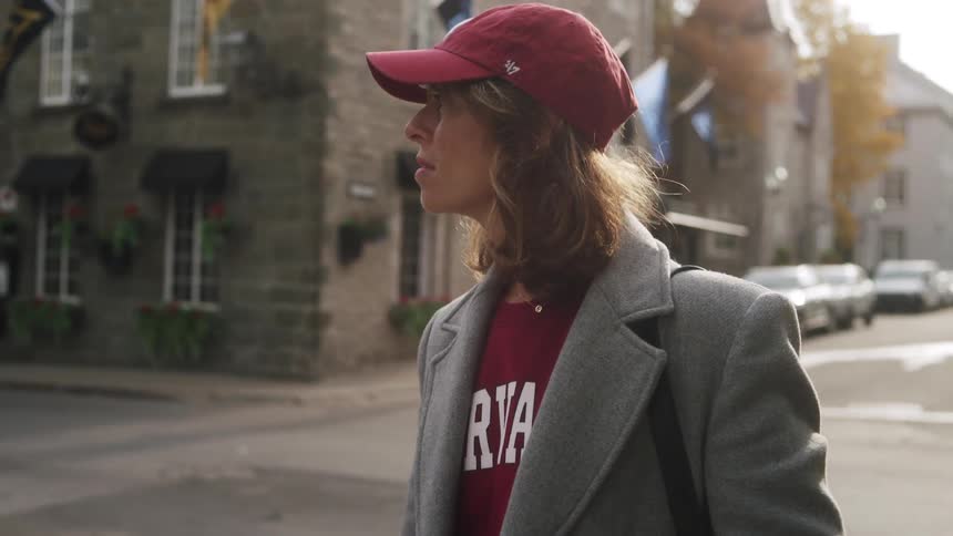A woman wearing a red baseball cap and a grey coat stands on a street corner looking around as a white van and a dark suv drive past her the scene is set in an urban area with stone buildings and blue flags visible in the background under bright dayligh