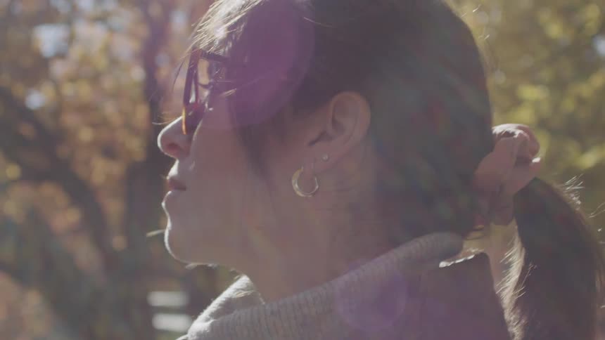 A woman wearing sunglasses gazes thoughtfully into the distance as warm sunlight filters through autumn trees behind her