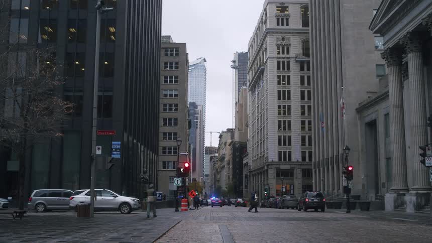 A narrow cobblestone street in a historic city center is framed by tall contrasting office buildings and classical architecture under a gloomy overcast skyin the distance police vehicles with flashing blue and red lights are parked at a crossroads