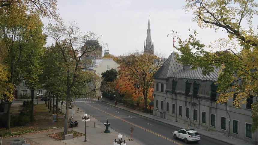 A white car travels down a quiet treelined city street bordered by historic stone buildings and autumn foliage in the distance the sharp spire of a cathedral and the iconic turrets of the fairmont le chteau frontenac rise against a pale sky