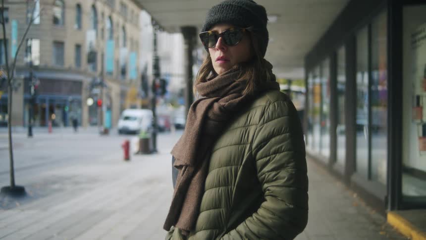 A woman dressed in a green puffer jacket brown scarf and black beanie stands on a city sidewalk while looking off to the side other pedestrians walk past her in the background as she remains still near a storefront