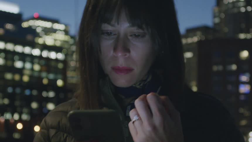 Woman standing on a downtown street using her phone as the light illuminates her face and lights from city buildings glow in the distance