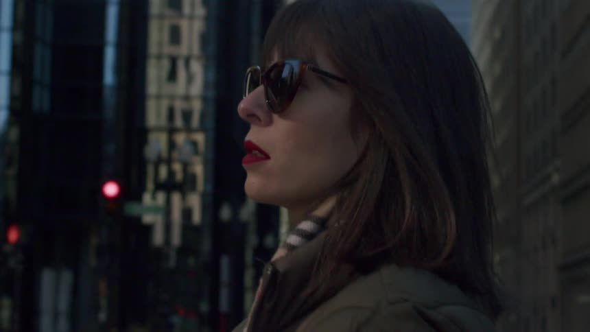 Woman wearing sunglasses and a coat crosses an intersection downtown during the daytime
