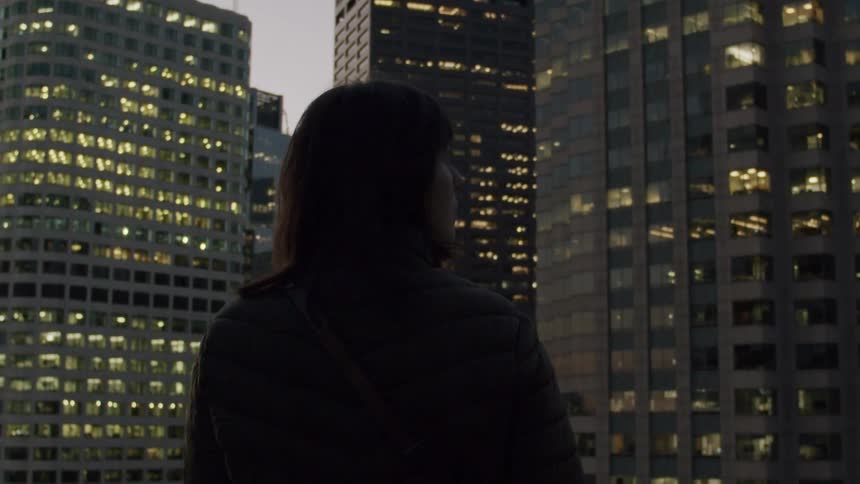 A woman is seen from behind looking out at a city skyline at dusk as she stands on a rooftop or high balcony the camera slowly zooms in on her and she turns her head slightly to the right to take in the view of the illuminated office buildings