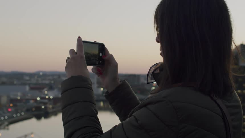 Woman captures an urban landscape at dusk using a smartphone from an elevated viewpoint