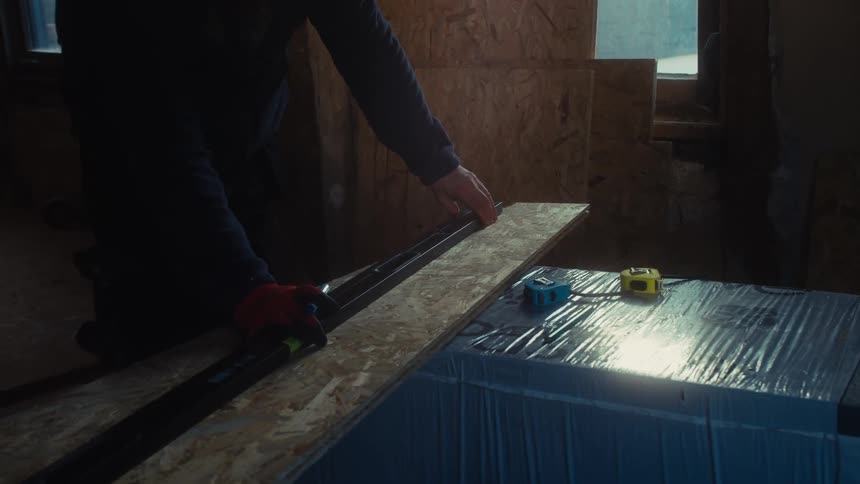 Person measures and marks a wooden panel in a construction setting with tools on the surface