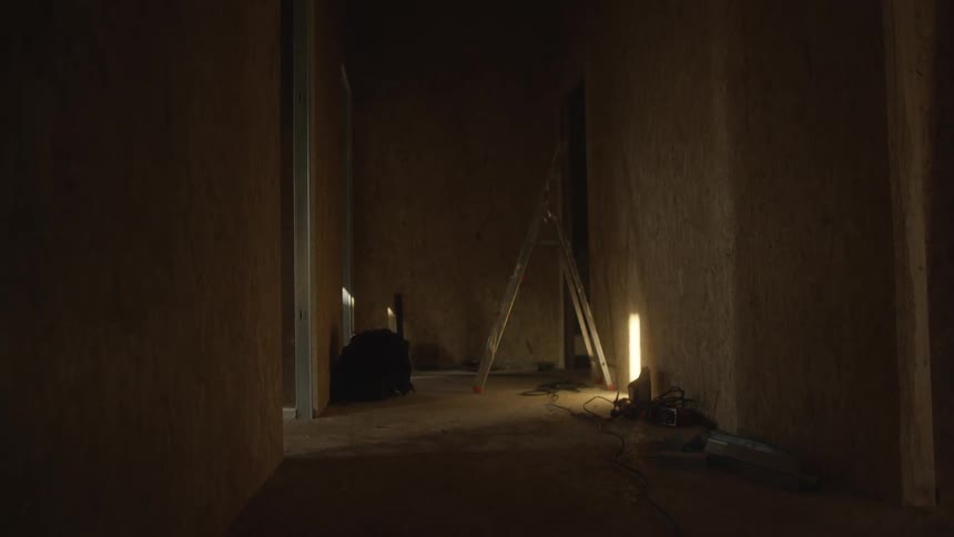The video shows a dimly lit narrow hallway or room under construction with rough unfinished walls a single bright work light stands on the floor casting long shadows and illuminating a silver ladder leaning against the back wall