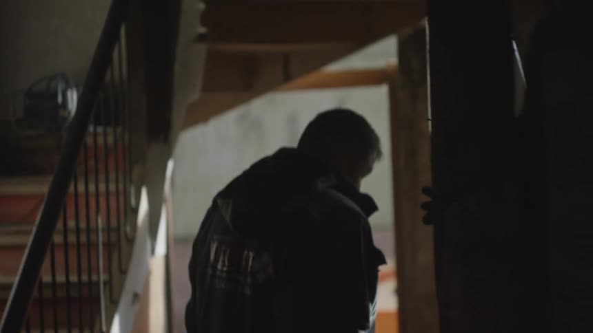 A man wearing a black jacket walks through a dark building and gets off the screen