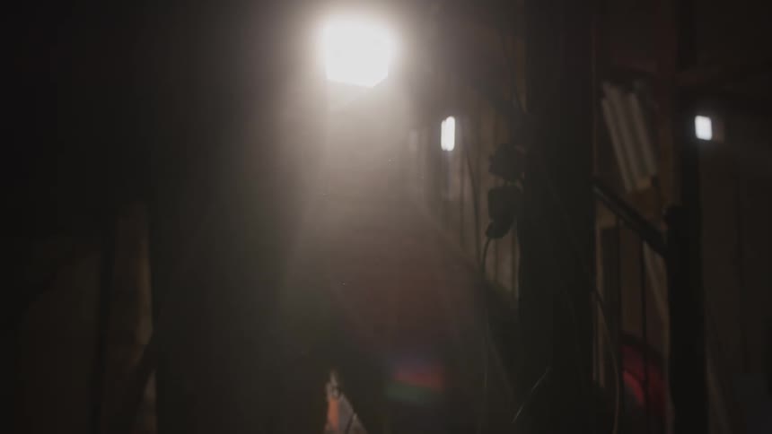 In a dimly lit attic or storage space a bright beam of sunlight streams through a small window illuminating swirling dust particles and creating a soft lens flare against the dark silhouetted interior