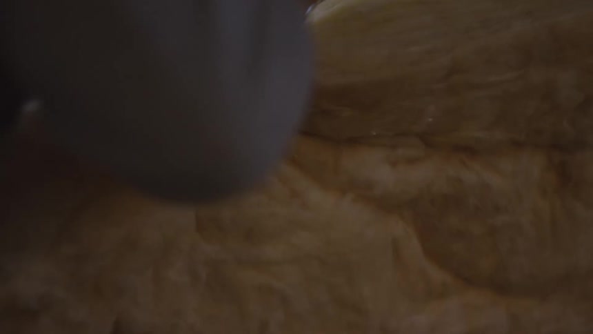 A worker wearing red gloves and a grey longsleeve shirt uses a utility knife to slice through a thick batt of yellow fiberglass insulation in a dimly lit indoor construction setting