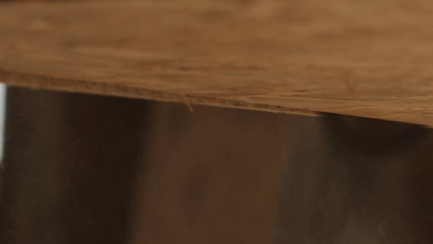 A circular saw blade cuts through a piece of wood from below sending dust and debris into the aira persons hand reaches down into the frame likely to move or adjust the material being worked on