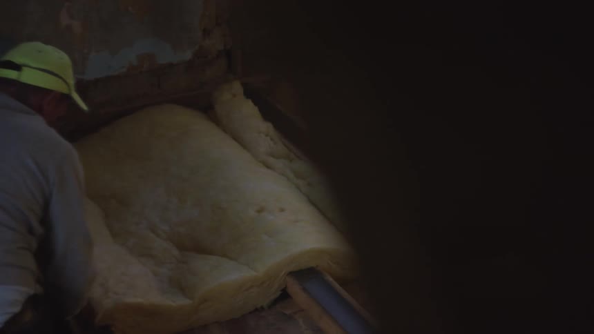 In a dimly lit and unfinished space a construction worker wearing a yellow cap installs a large roll of insulation into a wall