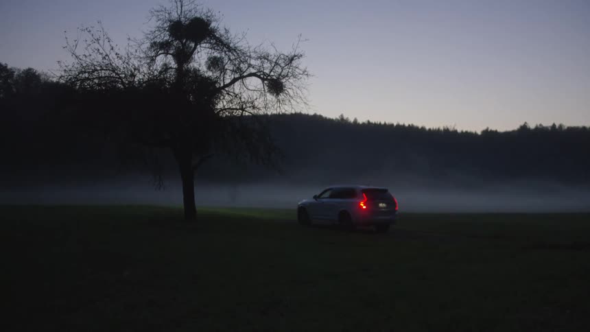 A white suv drives slowly across a dark foggy field during twilight passing behind a large silhouetted tree the cars headlights illuminate the misty grass while its red taillights remain visible as it moves toward the distant forested hills