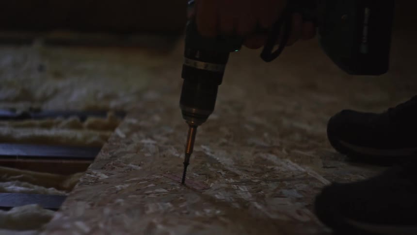 A person uses a cordless power drill to drive a screw into an osb board securing the subfloor to wooden joists above exposed fiberglass insulation in a dimly lit indoor construction site