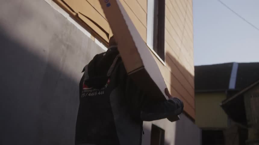 A worker carries a large cardboard geberit box on his shoulder while walking past a multistory buildingthe person is wearing a dark jacket with a logo and phone number printed on the back as they move through a narrow outdoor area