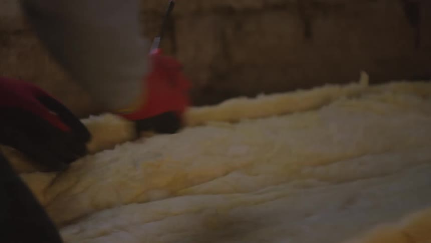 A person wearing red and black work gloves is shown carefully installing yellow fiberglass insulation between wooden floor joists