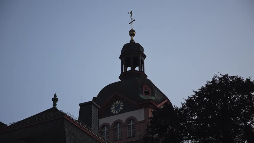 A church tower featuring a clock and a copper dome against a dim sky while the silhouettes of a nearby building and a leafy tree frame the architectural structure in the fading light