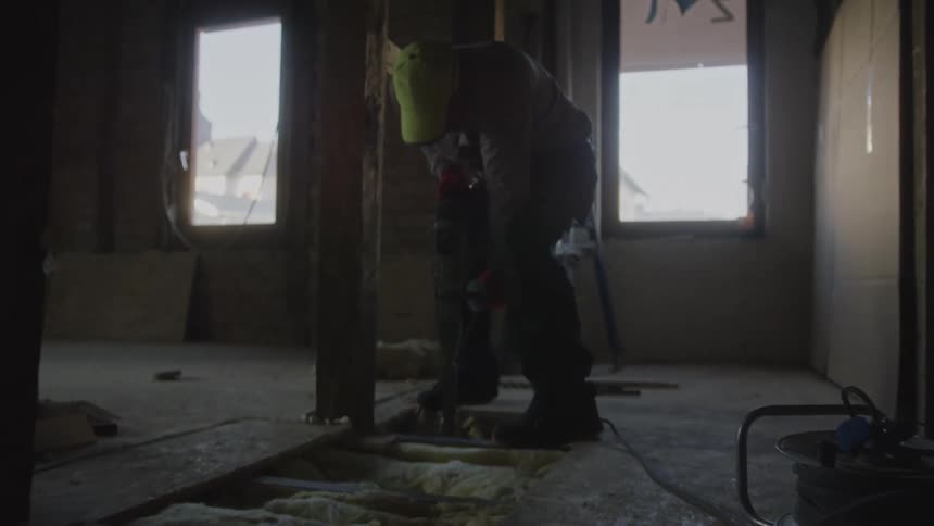 Construction worker drilling against the wooden floor of an unfinished building indoors during day time