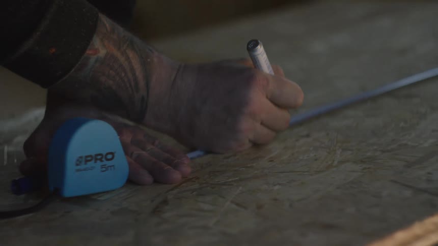 Male hands on a workshop using a measuring wheel to draw marks on a wooden board indoors during day time