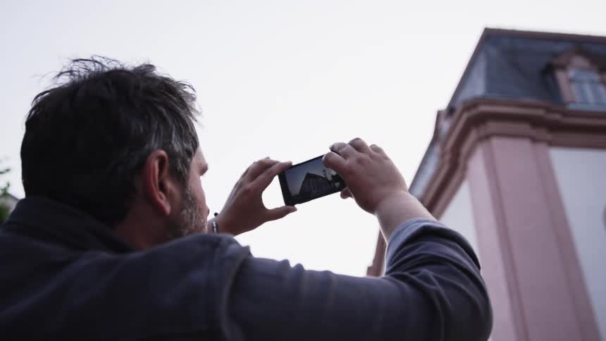 A man with dark hair stands with his back to the camera holding up a smartphone to photograph a large buildingthe building features a pale pink facade with classical architectural details and large arched windows under a grey roof