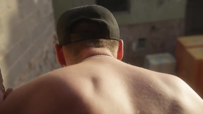 The person is seen from the back showing their bare neck and shoulders while wearing a dark baseball cap and a thin silver chainthe scene is set outdoors in bright sunlight with a stone wall to the left and several yellow crates or boxes stacked in t