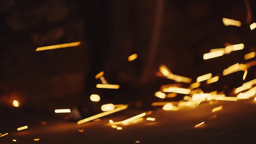 Bright orange and yellow sparks fly horizontally across a dark industrial background creating long trails of light as they collide with the ground