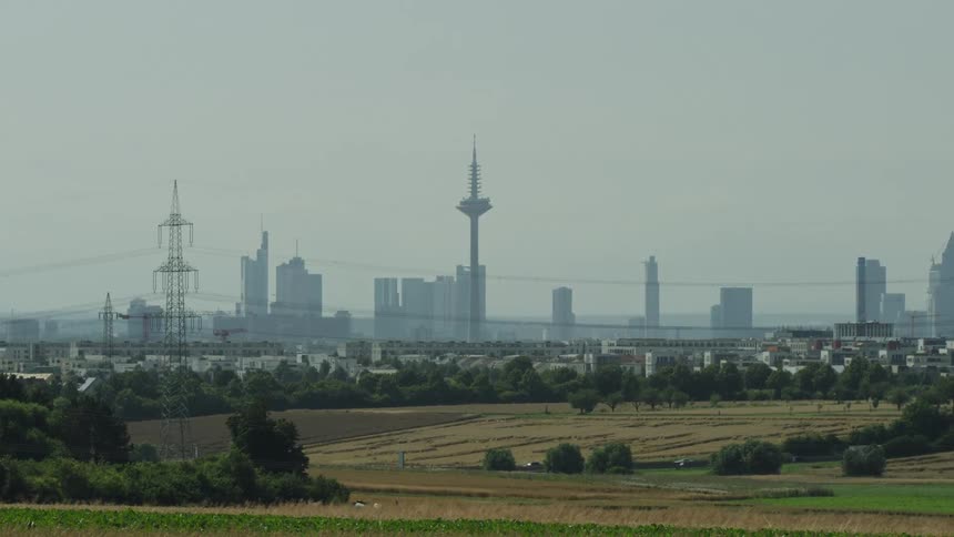 A distant city skyline with several skyscrapers and a tall communications tower sits under a hazy sky in the foreground green fields and power lines lead toward the urban center
