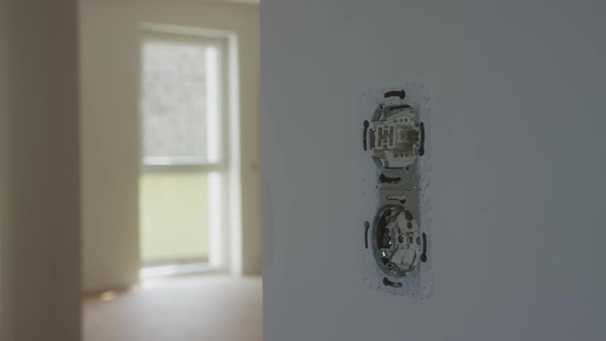 A closeup shot shows a double electrical socket box installed into a white wall within a blurred interior room