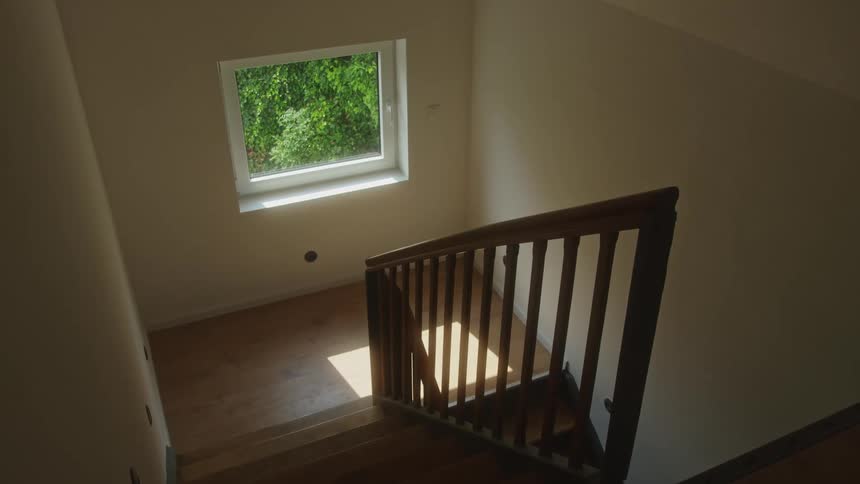 A downward view shows a wooden staircase and landing in a brightly lit home where sunlight streams through a square window overlooking green trees and casts linear shadows from the railing onto the floor