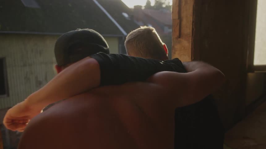 Two men stand with their arms around each others shoulders inside a derelict building looking out of a large window opening at a sundrenched neighborhood during sunset as the camera moves closer to them