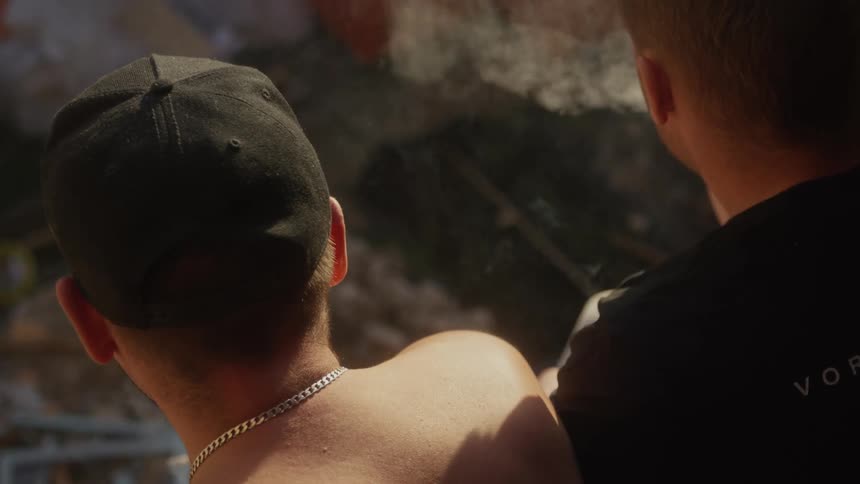 A silver or goldcolored curb chain necklace rests horizontally across the back of the neck of the individual on the leftthe brim of the black baseball cap creates a sharp curved line that separates the hat from the persons head and the background