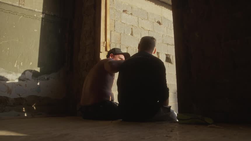 Two men sit on the floor of a derelict building and talk while looking out an open doorway into the sunlight