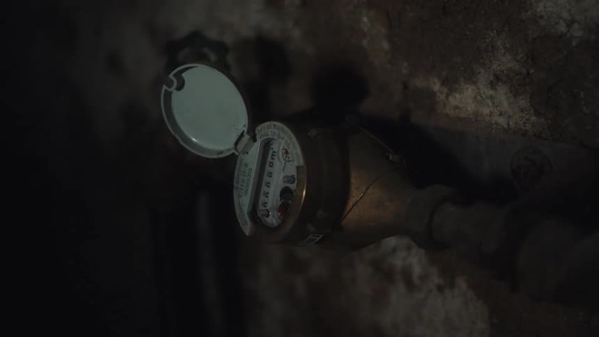 A closeup shot of a brass water meter mounted on a rough stone basement wall showing the mechanical counter and pipes while a flashlight beam intermittently illuminates the device in the dark room