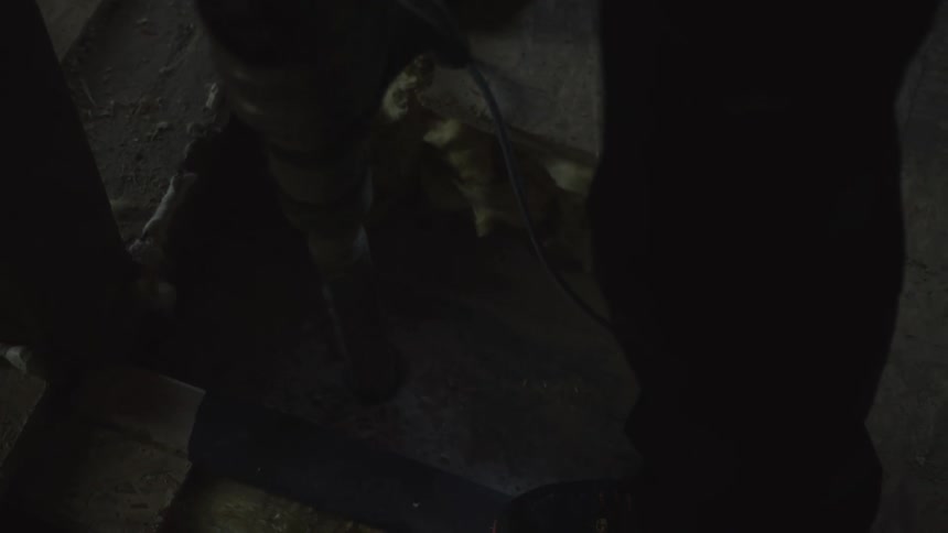 A construction worker uses a large power drill to bore a hole into a concrete floor inside a dark building frame near wooden studs and yellow insulation