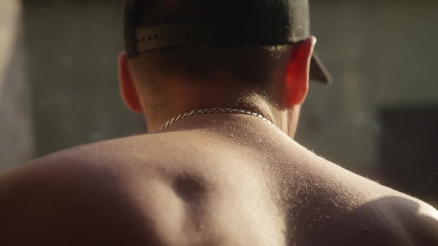 A man with a silver chain and black cap stands with his back to the camera his bare shoulders illuminated by warm sunlighthe appears to be exhaling smoke into the air while looking toward a blurred industrialstyle background
