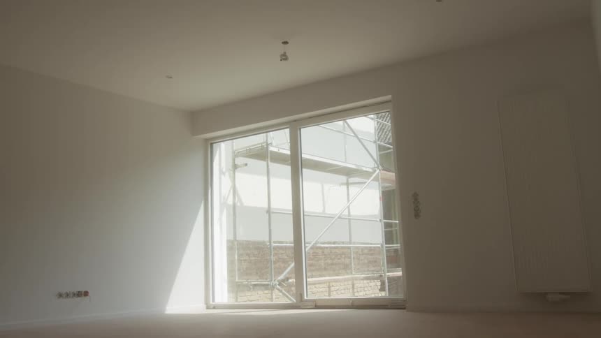 A bright empty room with white walls and a large glass door opening to a construction site outside
