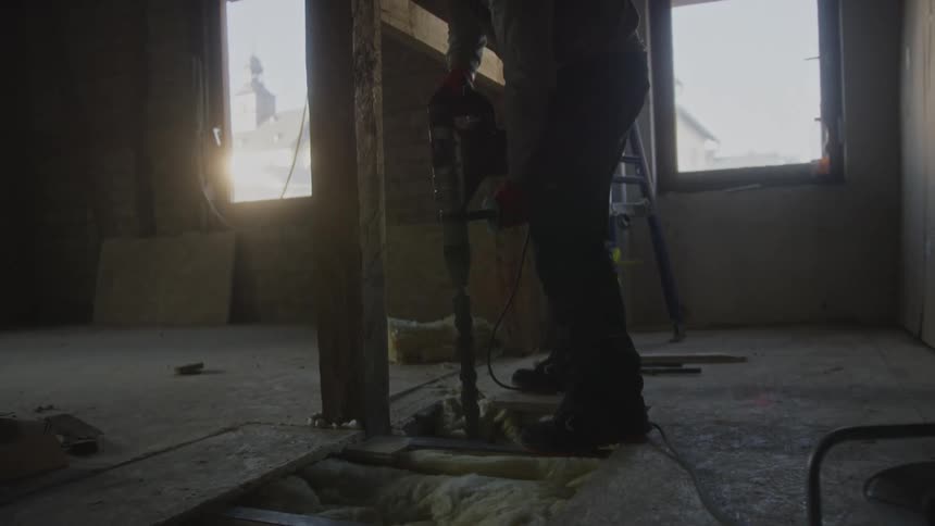 A construction worker in an attic uses a large power drill to bore into the wooden floorboards next to a support beam while sunlight streams through a nearby window