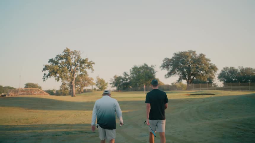 Two men are seen walking across a green golf course during the late afternoon here is a description of the scenetwo golfers walk sidebyside toward a distant hole on a sundrenched golf course carrying their clubs as the golden hour light casts long