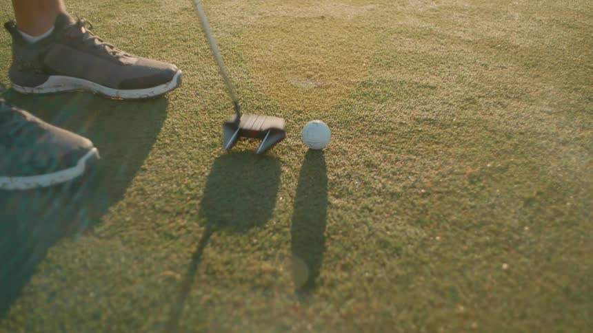 Male golf player rehearsing his swing with a golf club next to a ball outdoors during sunset