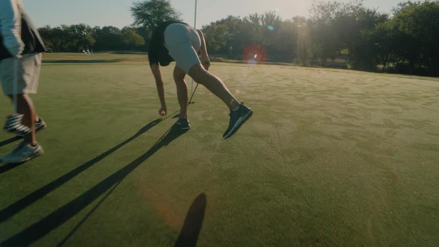 Two golfers walking on a sunlit green with one bending over to retrieve a ball near the flag