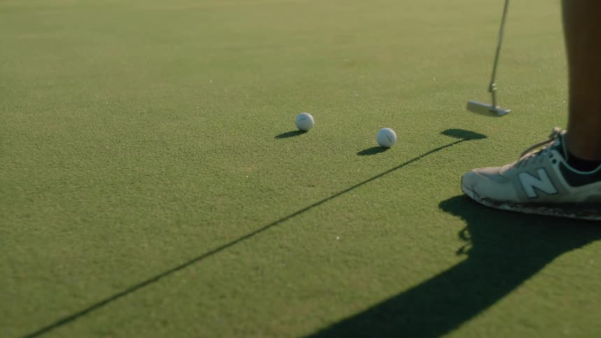 A golfer wearing new balance shoes stands on a putting green with a putter positioned near two golf balls the golfer strikes one of the balls sending it rolling across the short green grass