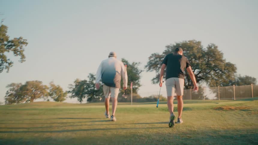 Two men walk into a grass covered field carrying golf clubs