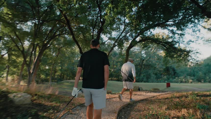 Two men carry golf clubs as they walk across a footbridge to an open field