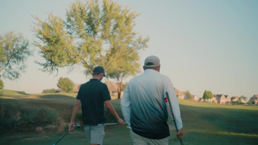 Two men carrying golf clubs walk across a sun drenched golf course