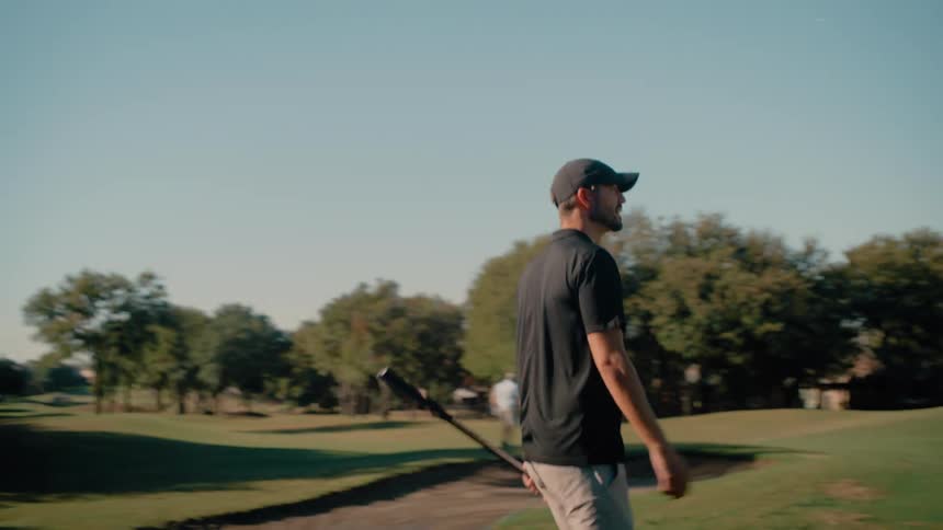 Golfer walks across the green holding a club with other players in the background